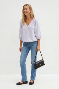 A smiling woman wearing a Naked Zebra - Elastic Puff Sleeve V-neck Blouse: Burgundy / L, blue jeans, and black ballet flats, carrying a black shoulder bag.