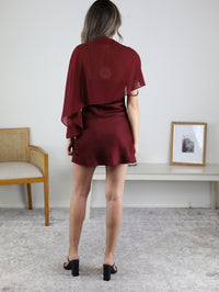A person seen from behind wearing an Amy Mini Dress with Sheer Cape in dark red satin with a matching sheer cape and black heeled sandals.