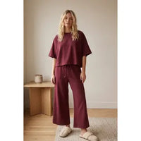 A woman stands indoors wearing the Heather Brushed Stretch Boxy Top & Wide Leg Pant Set in maroon, styled with cream slippers. She is on a light rug next to a wooden bench topped with a small vase, in a minimally decorated room.