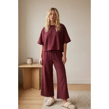 A woman stands indoors wearing the Heather Brushed Stretch Boxy Top & Wide Leg Pant Set in maroon, styled with cream slippers. She is on a light rug next to a wooden bench topped with a small vase, in a minimally decorated room.