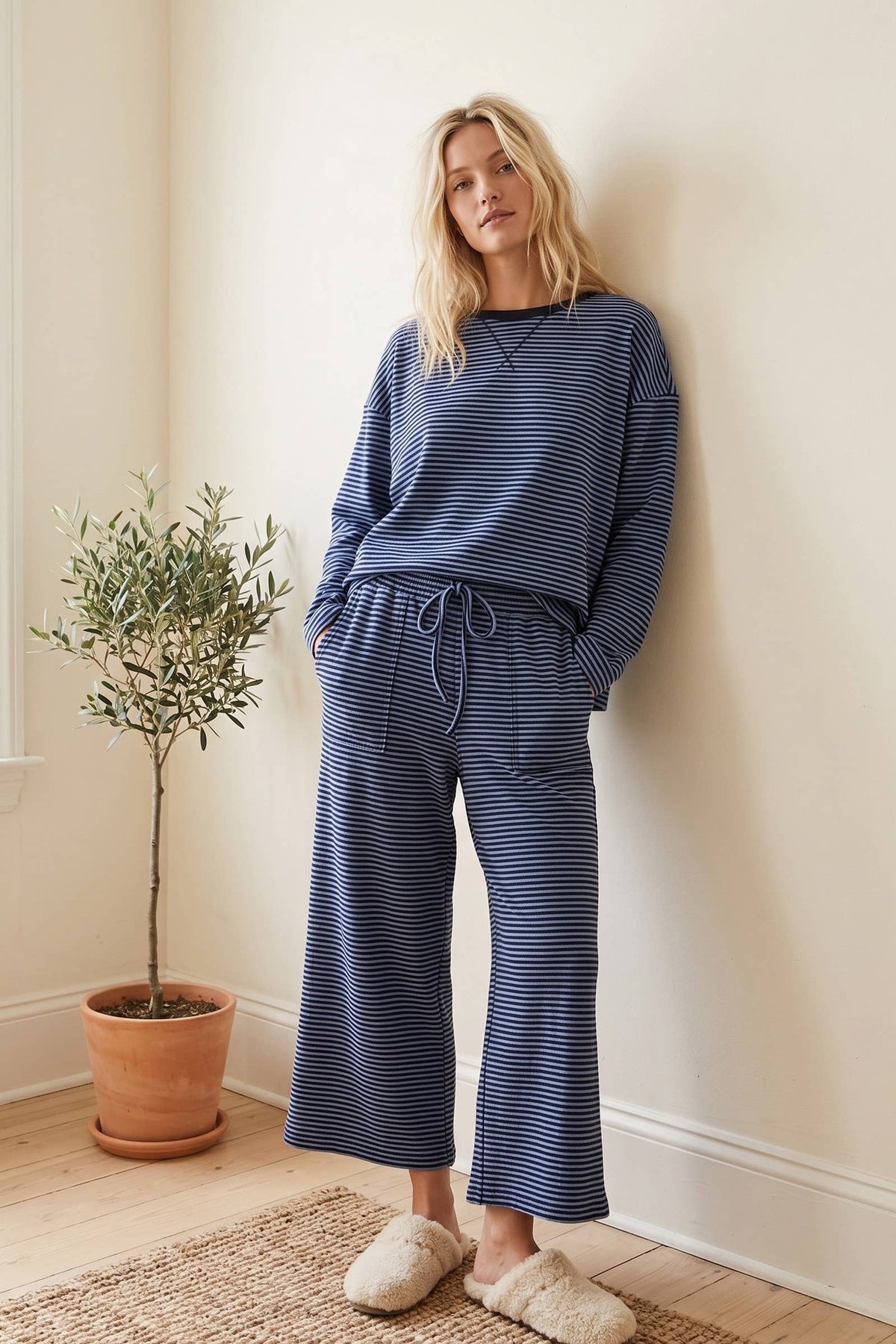 A person stands indoors by a potted plant, wearing the Misty Striped Relaxed Top & Wide Leg Pants with fuzzy slippers, hands in pockets, leaning against a beige wall.