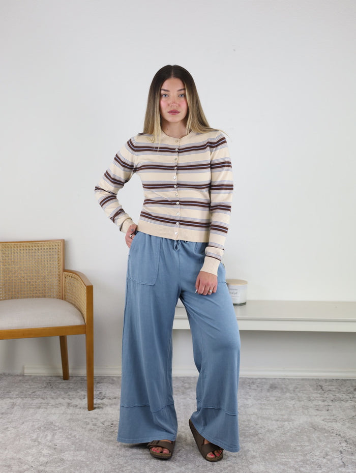 A woman stands indoors wearing the Sausalito Soft Striped Cardigan and loose blue pants, with one hand on her hip. A wooden chair and a white bench are visible in the background against a plain wall.