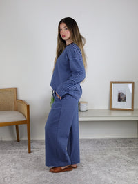 A person stands sideways in a Misty Striped Relaxed Top & Wide Leg Pants set—loose blue-and-white stripes, wide leg pants, and brown flats—in a simple room with a chair, shelf, candle, and framed photo.