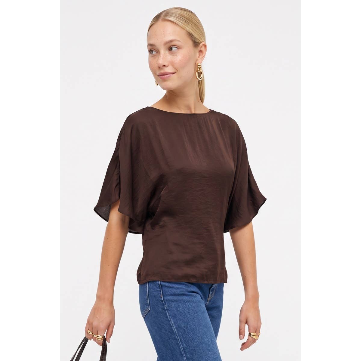 A woman wearing a Naked Zebra - Back Tie Dolman Sleeve Silky Fall Blouse in dark navy and blue jeans.