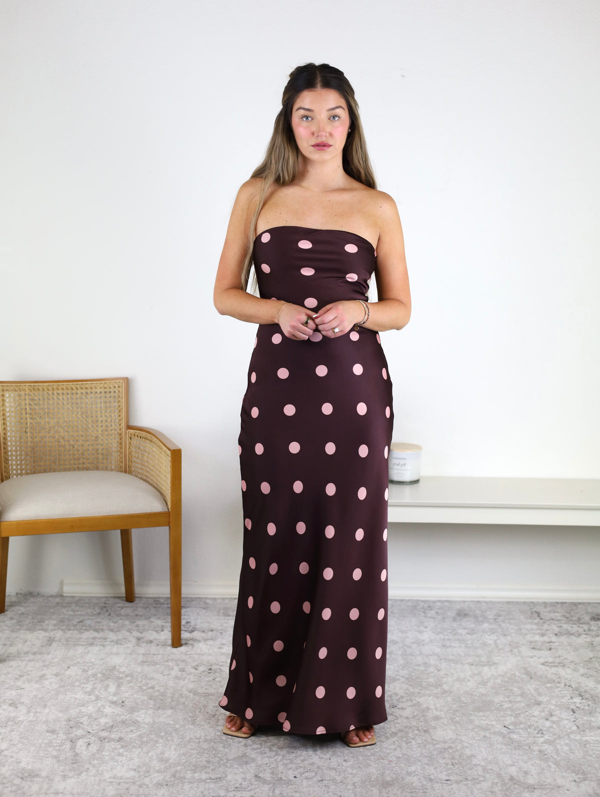 A woman stands indoors wearing the Polka Dot Satin Strapless Maxi Dress, a brown maxi dress adorned with large pink polka dots. She is barefoot on a gray carpet; behind her are a wooden chair and a white shelf against a plain white wall.
