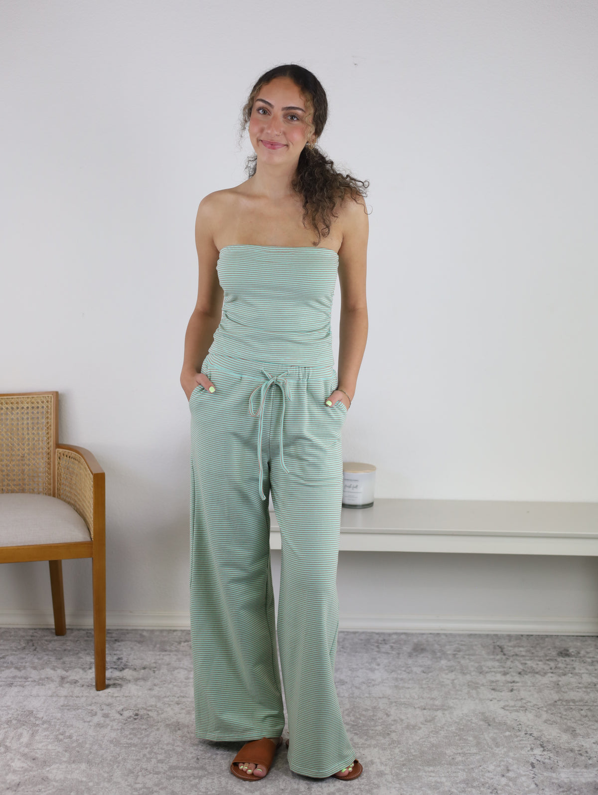 A woman stands indoors, smiling with her hands in her pockets, wearing the Kia Striped Tube Top and Pants Set—a stylish ensemble featuring a light green, strapless top and flowing women’s wide-leg pants. Behind her, a chair and bench rest on a light-colored rug.