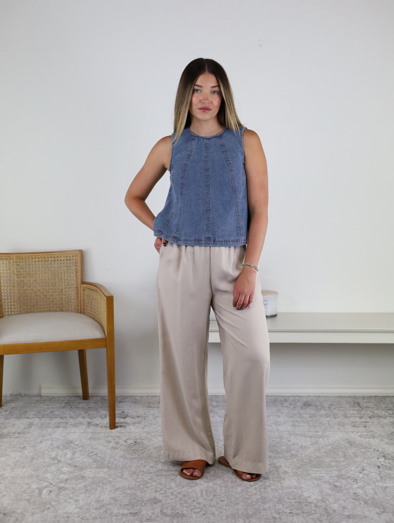 A woman stands indoors wearing the Rebecca Seam Detail Sleeveless Denim Top – Final Sale, styled as a versatile summer blouse with beige wide-leg pants and brown sandals. She poses with one hand on her hip in a minimal room featuring a chair and a bench against a white wall.