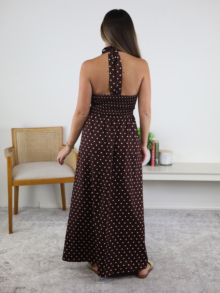 A woman stands indoors, facing away, wearing the Marley Polka Dot Halter Maxi Dress in brown with white polka dots. This vacation-ready dress features a sleeveless halter-neck design and flows elegantly to her ankles, making it perfect for resort wear. She is positioned on a light carpet near a wooden chair and a white shelf decorated with various decor items.