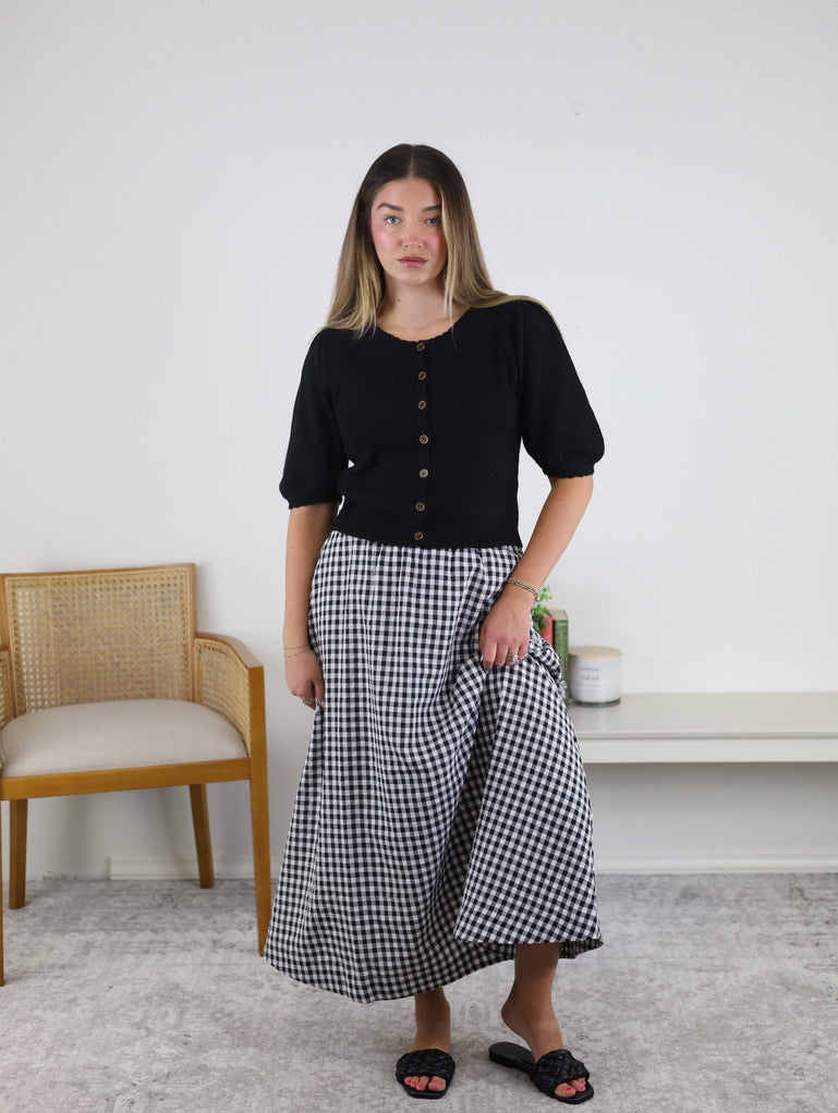 A woman stands indoors wearing the Monica Short Sleeve Cardigan in black, styled with a long black-and-white checkered skirt and black sandals. In the background, there is a wooden chair and a white console table adorned with decor items.