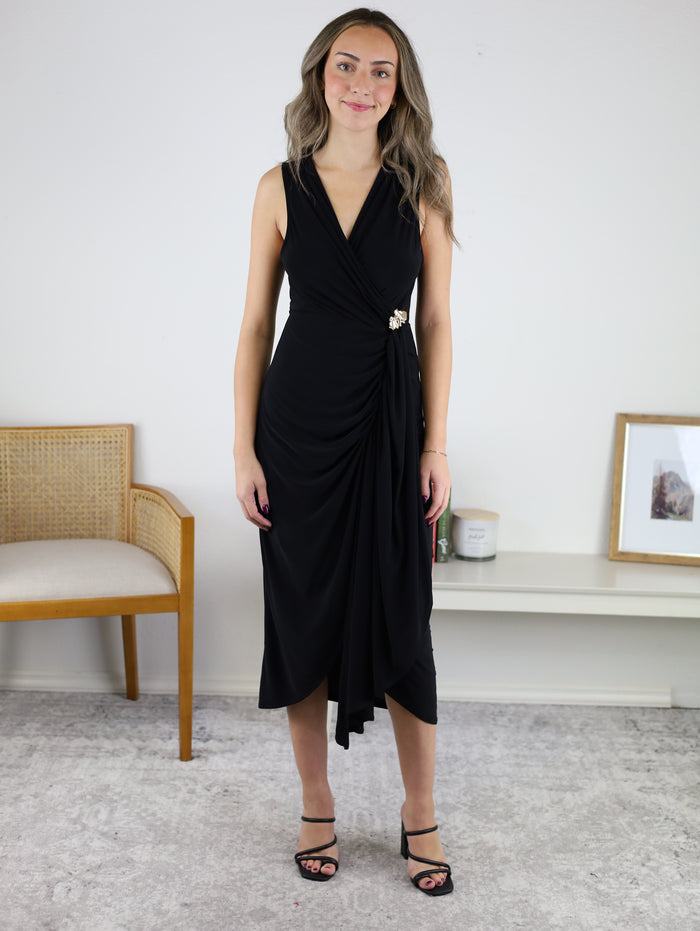A woman wears the black Kamden Side Gather Midi Dress, a sleeveless V-neck style featuring a side-gathered waist with decorative embellishment and an asymmetrical hemline.