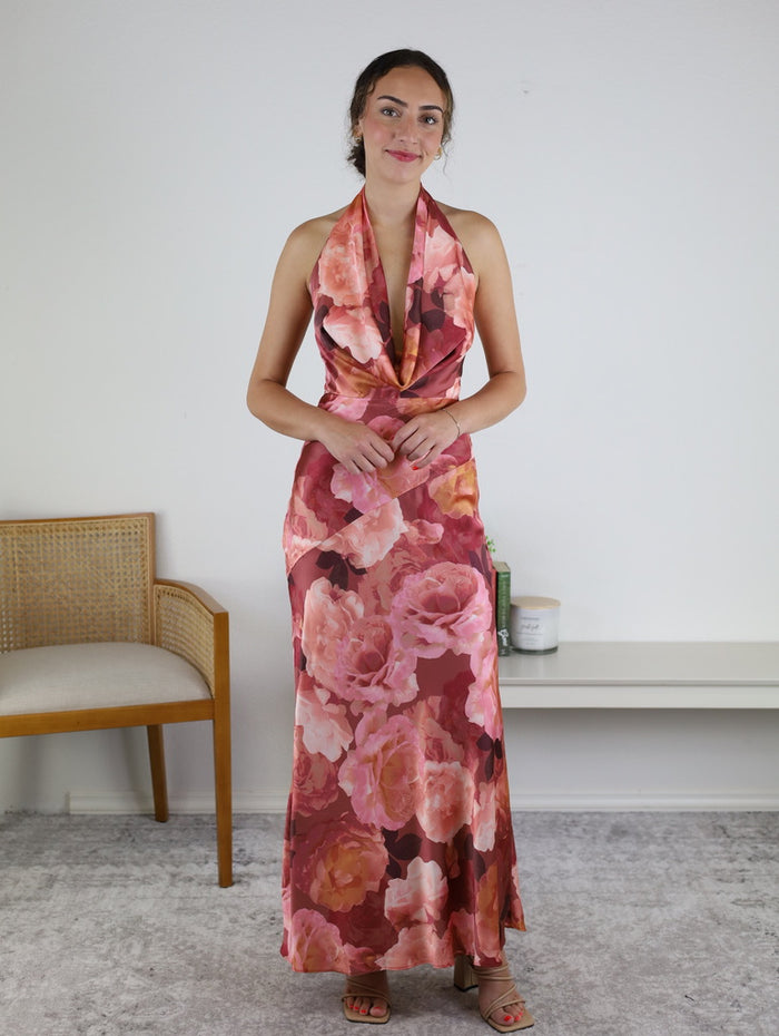 A woman stands indoors, smiling warmly with her brown hair tied back, wearing the Presley Floral Draped Halter Neck Maxi Dress—a stunning piece featuring vibrant pink and orange floral patterns. In the background, a wooden chair and a white shelf decorated with charming small accents complete the inviting scene.