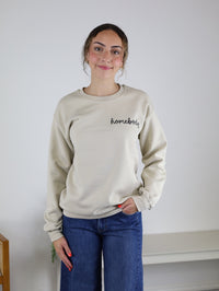 Wearing the Homebody Sweatshirt—a relaxed fit, beige unisex top crafted from a soft cotton polyester blend and featuring “homebody” lettering—a person with curly hair pairs it with blue jeans while standing and smiling in a simple, light-colored room.