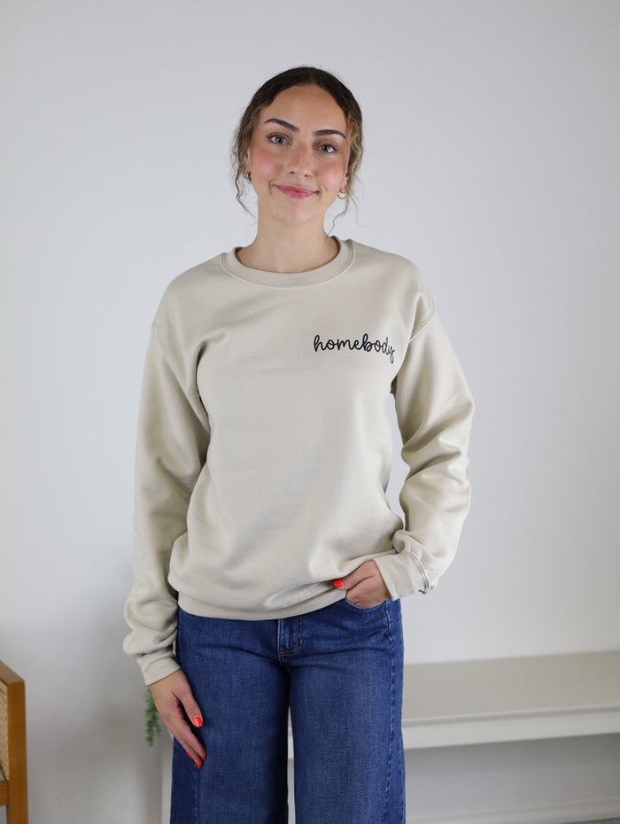 Wearing the Homebody Sweatshirt—a relaxed fit, beige unisex top crafted from a soft cotton polyester blend and featuring “homebody” lettering—a person with curly hair pairs it with blue jeans while standing and smiling in a simple, light-colored room.