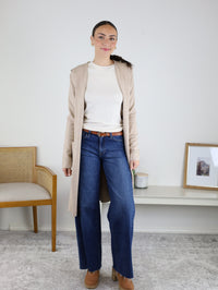 Wearing the Pinch Me Long Cardigan in beige, a woman stands indoors layered over a white sweater with blue wide-leg jeans and brown shoes. Set against a white wall with a chair and framed picture in the background, this look is perfect for cozy layering on cooler days.