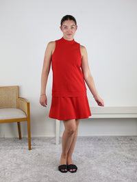 A woman stands indoors on a light gray carpet, wearing the Cooper Sleeveless Mock Neck Shift Dress in red. She is barefoot, holding black sandals, and poses with a relaxed expression. In the background are a beige chair and a white bench.