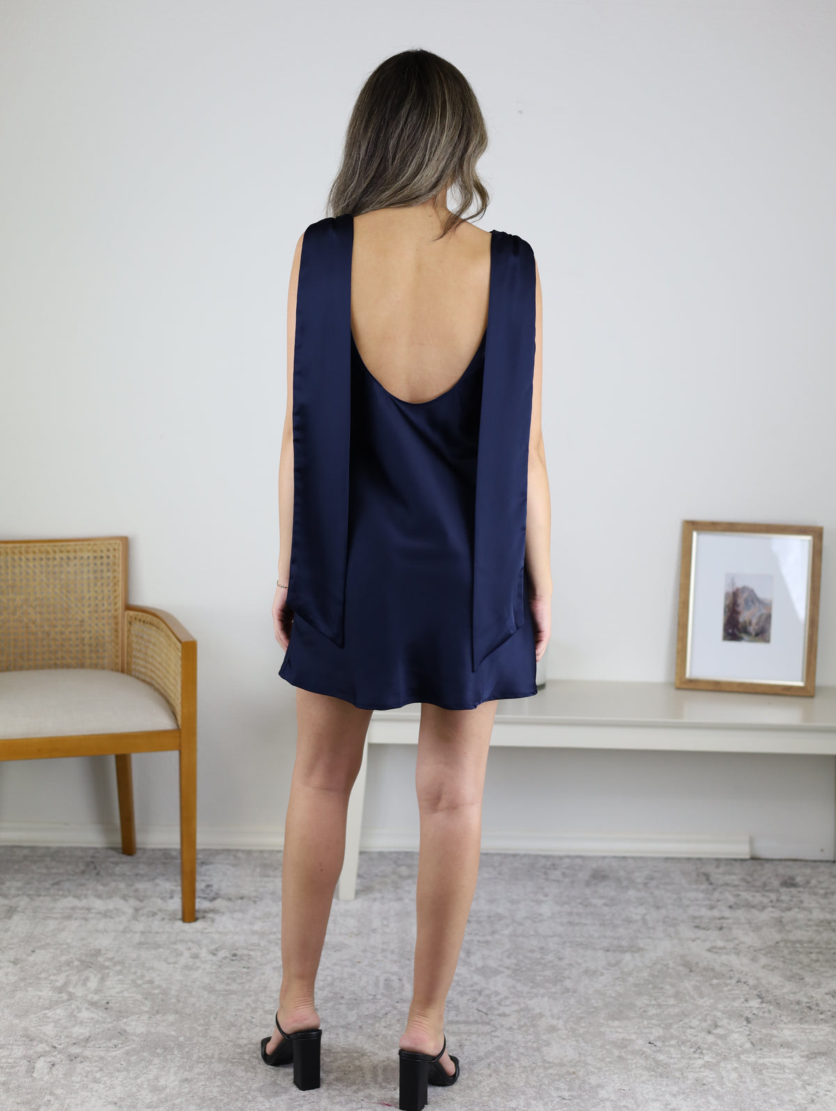 Back view of a person wearing a navy blue satin Hanford Cowl Neck W/Scarf Mini Dress with a low-cut back, wide shoulder straps, and black heeled sandals.