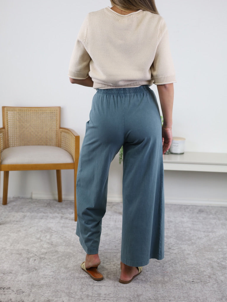 A person stands indoors facing away, wearing a beige short-sleeve top, the Scout Jersey Crop Flare Pant in Emerald Cove with cropped flare legs, and tan flat shoes. A wooden chair and a white table are in the background on a light-colored carpet.