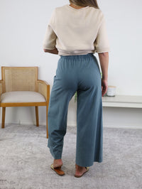 A person stands indoors facing away, wearing a beige short-sleeve top, the Scout Jersey Crop Flare Pant in Emerald Cove with cropped flare legs, and tan flat shoes. A wooden chair and a white table are in the background on a light-colored carpet.
