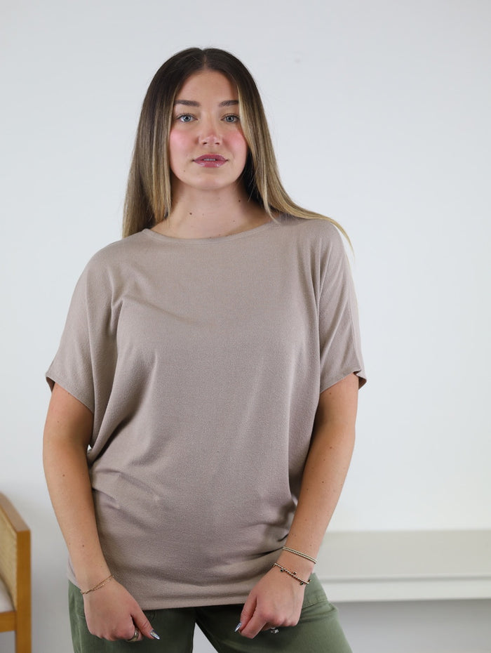 A woman with long brown hair wears the loose, taupe Wallace Top with dolman sleeves, paired with green pants. She stands against a plain white wall with part of a chair visible on the left, looking forward with a neutral expression.