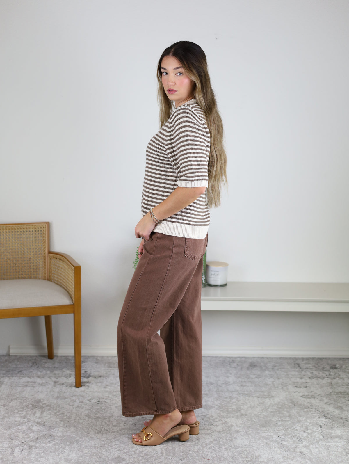 A woman with long hair stands sideways, wearing the Scout Striped Sweater, wide brown pants, and sandals. A wooden chair and a white shelf with a candle are in the background of the minimal room.