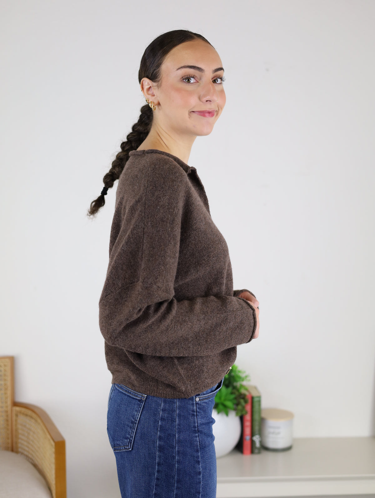 A woman with dark braided hair, wearing the Beth Lightweight Cardigan and blue jeans, stands sideways and smiles. A table with books, a candle, and a plant is in the background.