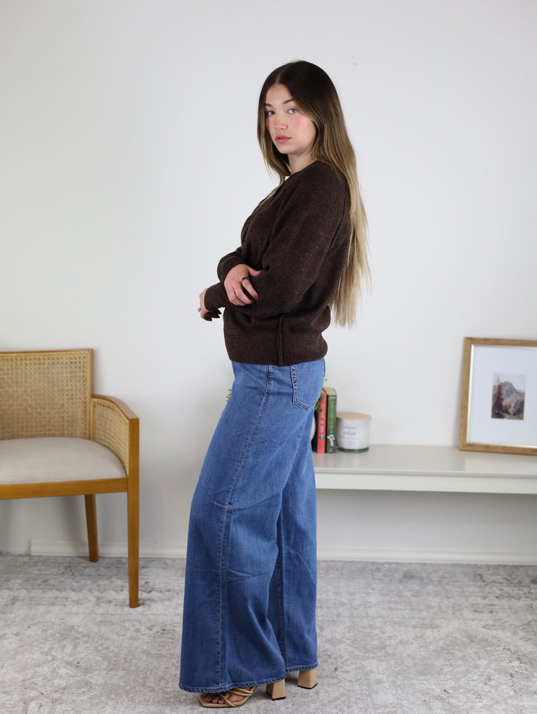A woman with long hair wears the Theo Sweater and wide-leg blue jeans, standing sideways in a minimalist room with a chair, low shelf, books, and framed artwork on a white wall.