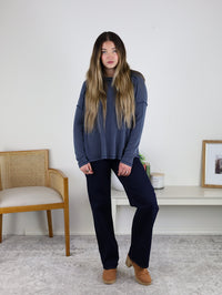A woman with long hair stands indoors in a loose blue ribbed knit Finley Sweater, dark pants, and tan shoes beside a wooden chair and a table with decor, against a white wall.