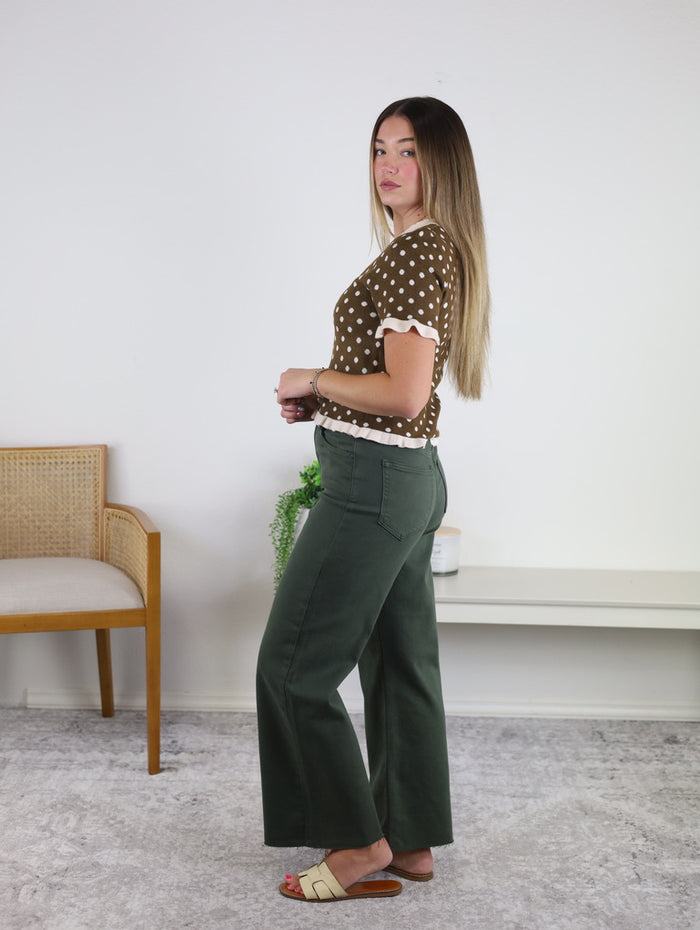 Wearing the Lainey Polka Dot Sweater, a woman with long straight hair pairs it with green wide-leg pants and sandals as she stands sideways in a minimalist room furnished with a chair, plant, and bench.