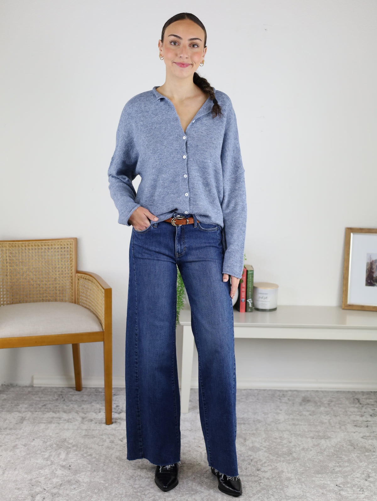 A woman stands indoors, smiling with one hand in her pocket, wearing the Beth Lightweight Cardigan in blue, paired with wide-leg jeans and black shoes. A wooden chair and a shelf filled with books and decor are visible behind her.