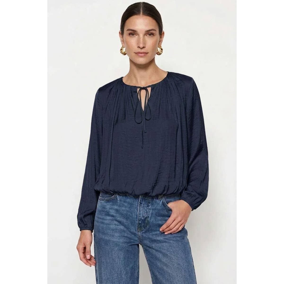 A woman wears a Naked Zebra - Long Sleeve Draped Bubble Hem Blouse in wine, featuring a gathered neckline, tie detail, and bubble hem, paired with blue jeans.