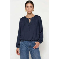 A woman wears a Naked Zebra - Long Sleeve Draped Bubble Hem Blouse in wine, featuring a gathered neckline, tie detail, and bubble hem, paired with blue jeans.