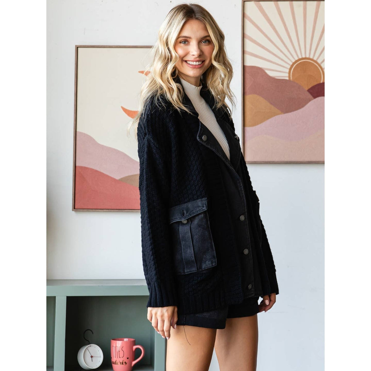 A smiling woman indoors wears the Veveret - Knit Body Denim Layerd Jacket: Cream / Denim / L. Colorful abstract mountain art prints, a pink mug, and decor are visible on a green shelf behind her.