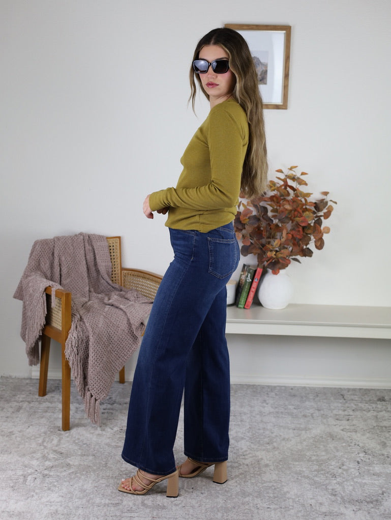 A woman wearing a long-sleeved, ribbed Dijon yellow Dee Ribbed Round Neck Tee, dark blue wide-leg jeans, and tan heeled sandals.