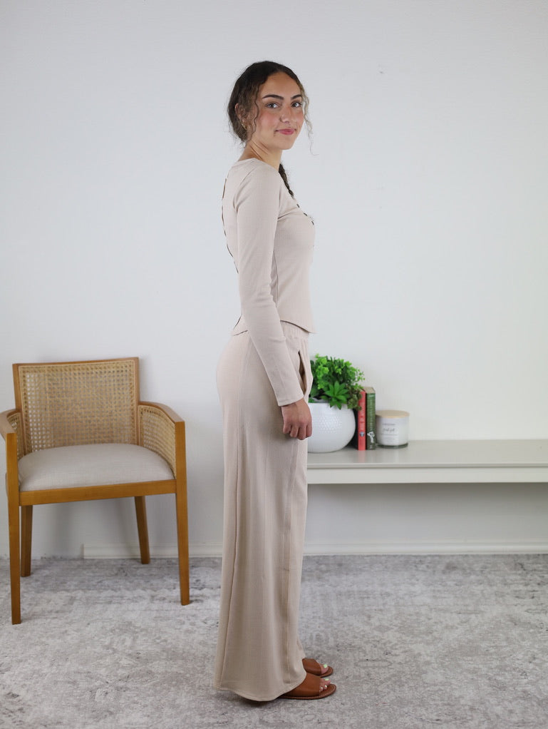 A woman stands sideways wearing the Statler Lounge Set, which features a beige long-sleeve henley top and matching wide-leg pants, styled with brown sandals. In the background are a wooden chair and a shelf with books and a plant.