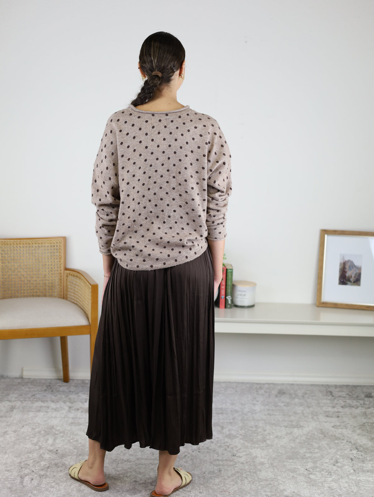 A person with braided hair stands facing away, wearing a tan polka-dot sweater paired with the Maci Satin Crinkle Pleated Maxi Skirt—an ideal addition to any fall wardrobe. Sandals complete the look. There is a chair, a low shelf with decor, and a framed picture on the white wall.