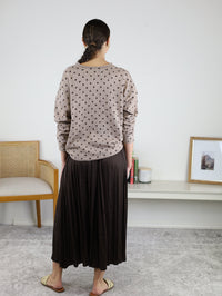 A person with braided hair stands facing away, wearing a tan polka-dot sweater paired with the Maci Satin Crinkle Pleated Maxi Skirt—an ideal addition to any fall wardrobe. Sandals complete the look. There is a chair, a low shelf with decor, and a framed picture on the white wall.