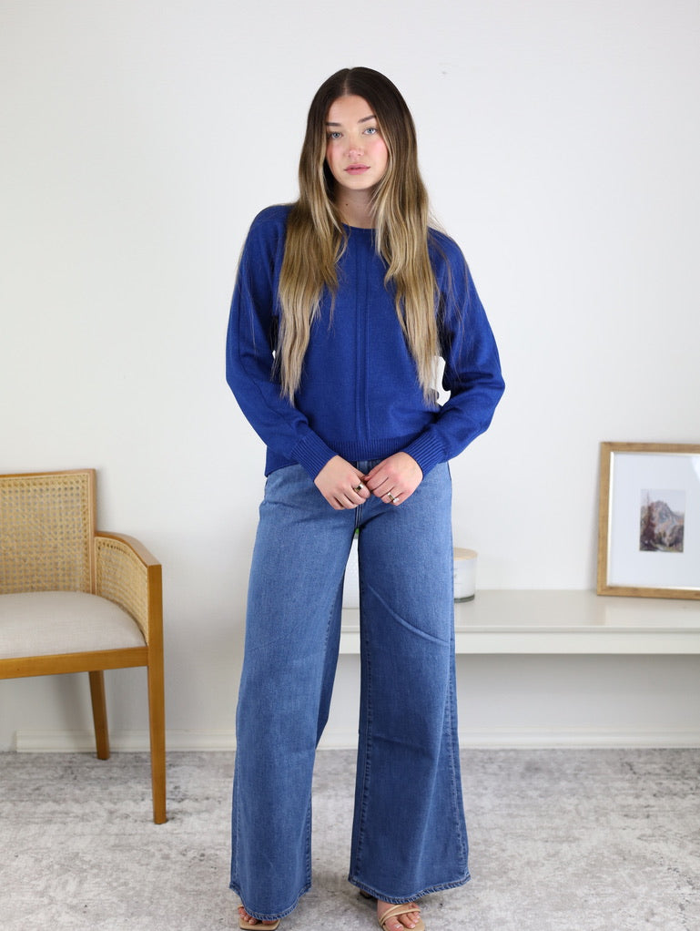 A person wearing an Eli Front Seam Sweater in royal blue viscose blend knit with a vertical front seam and wide-leg blue jeans.