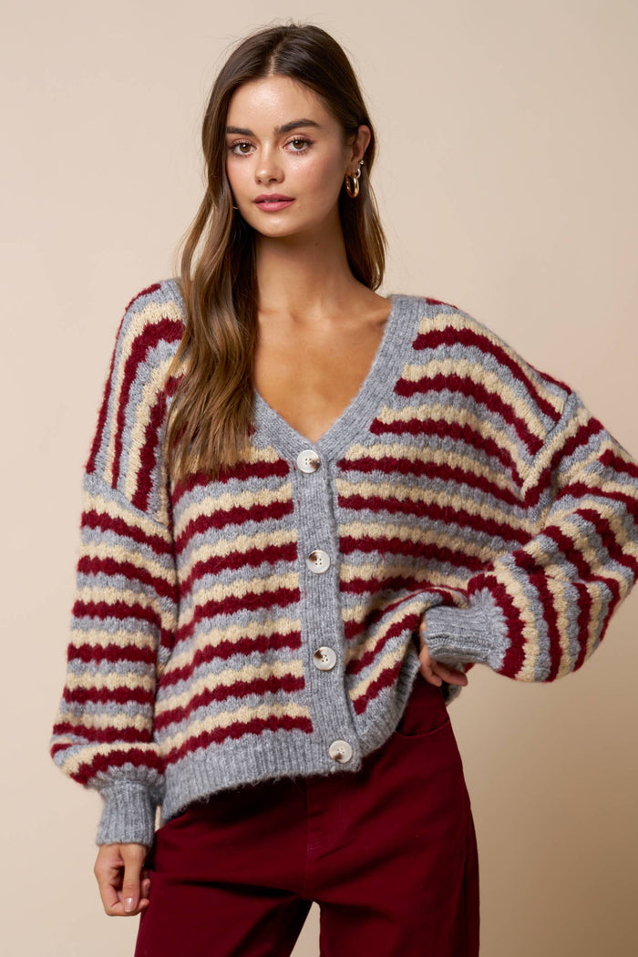 A woman stands against a beige background wearing the Lalavon Relaxed Stripe V-Neck Button Cardigan in grey, burgundy, and beige, featuring a loose fit, long sleeves, horizontal stripes in red and beige on gray fabric, and large white buttons. She pairs it with maroon pants.