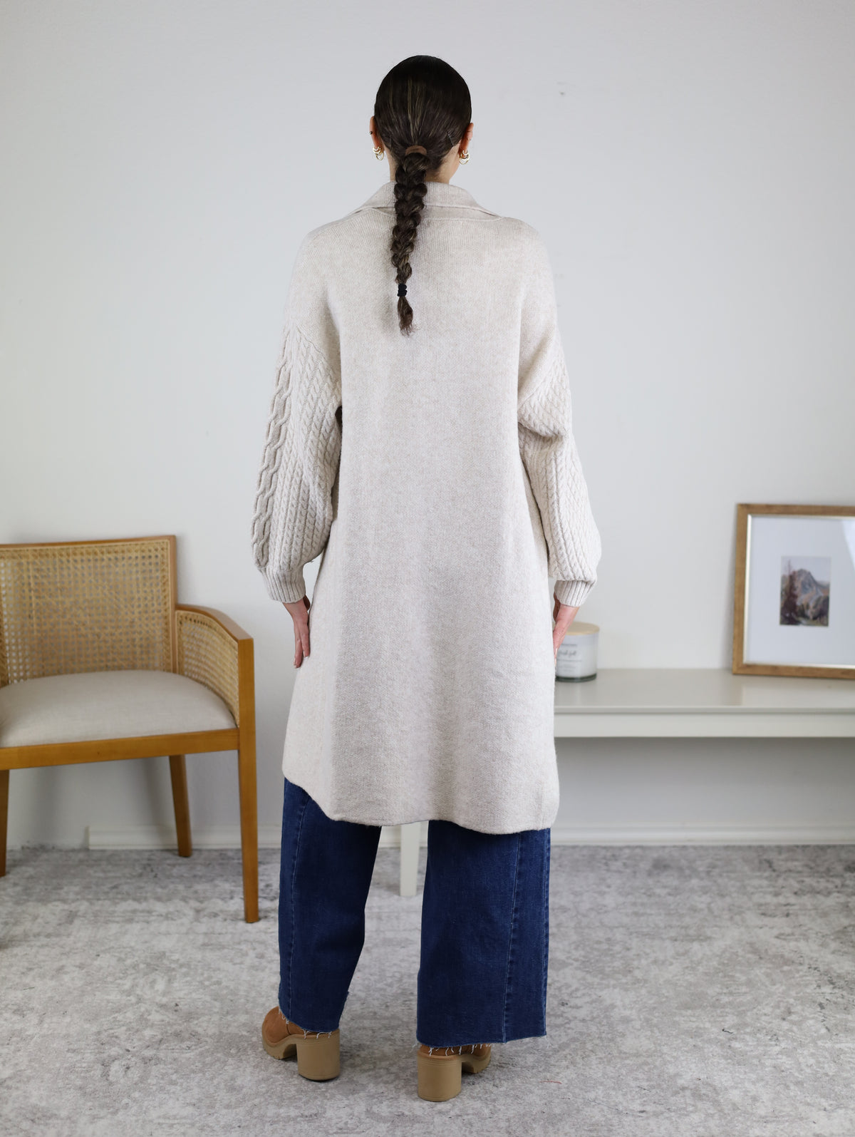 A person with a braid wears the Ashford Cable Knit Long Sleeve Knit Coat—a long beige sweater coat with textured sleeves—paired with wide-leg blue jeans and platform shoes, standing indoors facing away from the camera. A chair and framed art are visible in the background.
