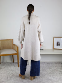 A person with a braid wears the Ashford Cable Knit Long Sleeve Knit Coat—a long beige sweater coat with textured sleeves—paired with wide-leg blue jeans and platform shoes, standing indoors facing away from the camera. A chair and framed art are visible in the background.
