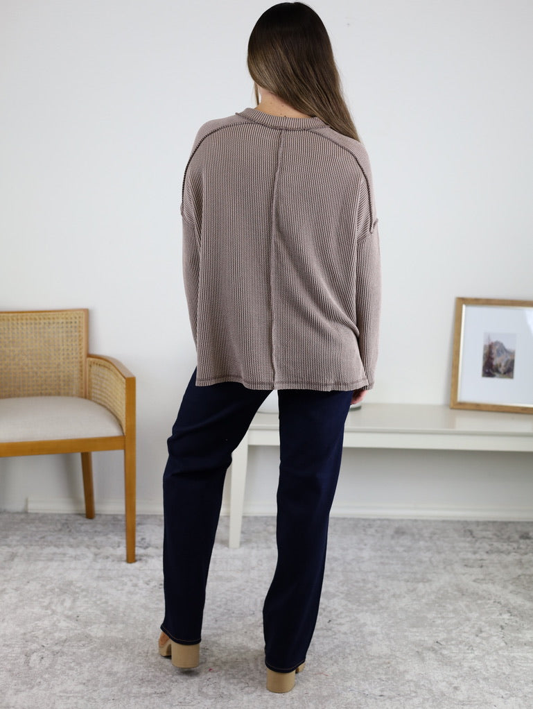 A person with long brown hair stands indoors, facing away, wearing a loose Finley Sweater with a ribbed knit texture, dark pants, and beige shoes. A wooden chair and white table are visible behind them.