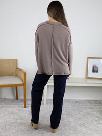 A person with long brown hair stands indoors, facing away, wearing a loose Finley Sweater with a ribbed knit texture, dark pants, and beige shoes. A wooden chair and white table are visible behind them.