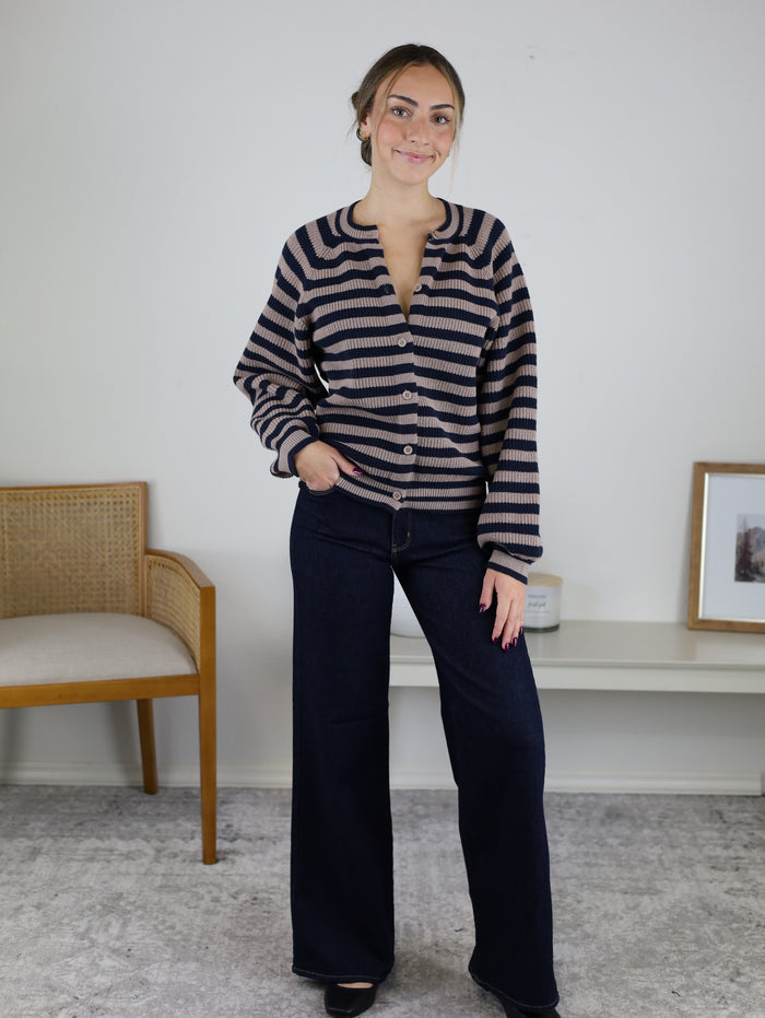 A woman wearing a brown and navy blue striped Justine Longsleeve Crewneck Striped Sweater with long sleeves, a button front, and wide-leg dark wash jeans.