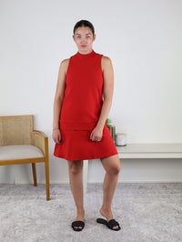 A woman in the Cooper Sleeveless Mock Neck Shift Dress stands indoors on a light carpet, next to a wooden chair and a white console table adorned with a candle and a small green plant. She is wearing black slide sandals.