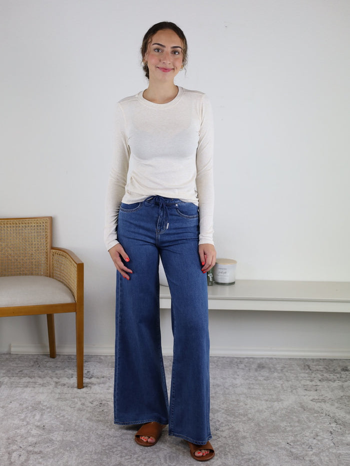 A woman stands indoors on a light gray carpet, wearing a long-sleeved white top and the Rain Mid Rise Wide Leg jeans made from breathable denim, paired with brown sandals. A wooden chair and a white shelf with candles are visible in the background.