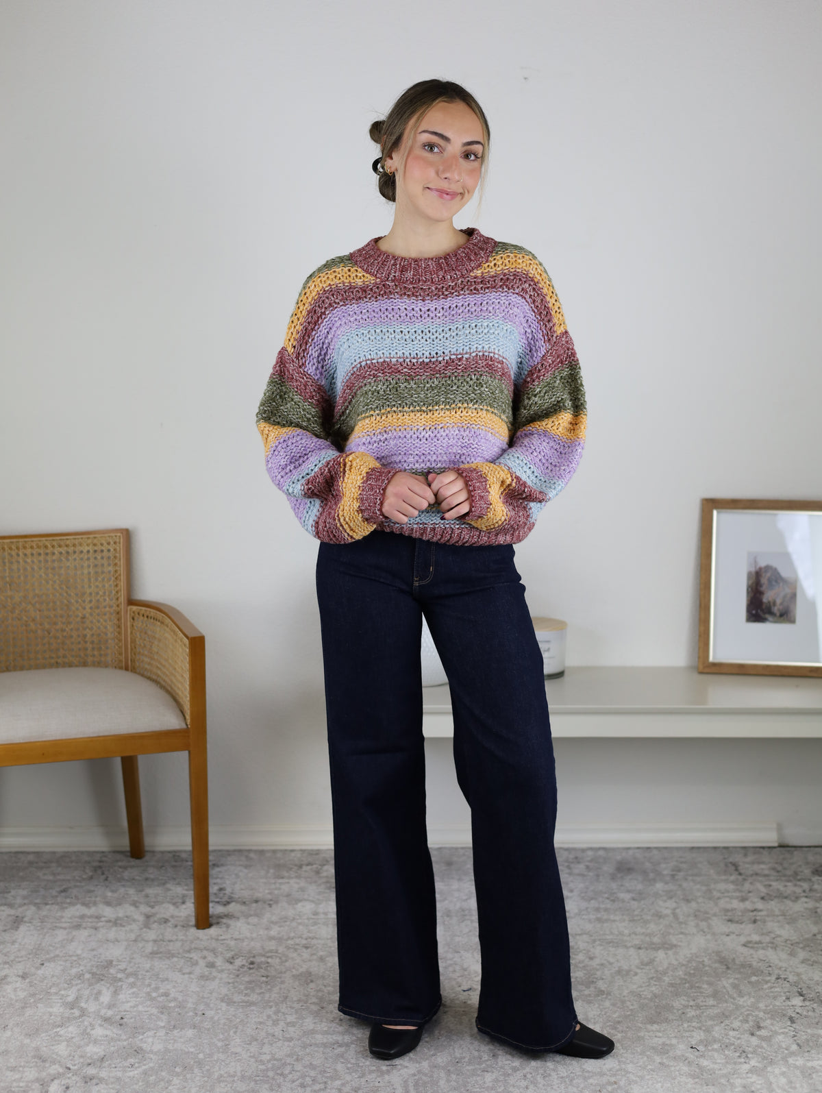 A woman wearing a cozy Reign Striped Knit Sweater in pink, lavender, olive green, and yellow, paired with dark wash wide-leg jeans.