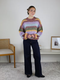 A woman wearing a cozy Reign Striped Knit Sweater in pink, lavender, olive green, and yellow, paired with dark wash wide-leg jeans.
