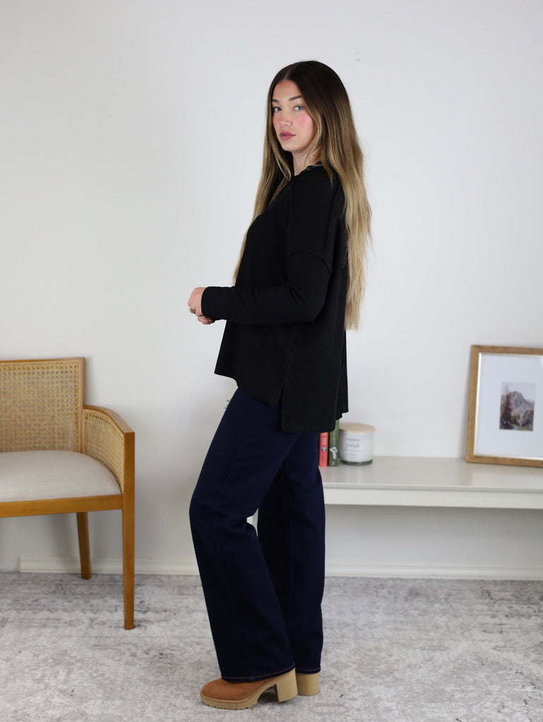 A woman with long hair stands sideways in a minimalist room, wearing the Finley Sweater, dark blue jeans, and tan platform shoes.