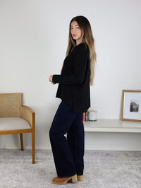 A woman with long hair stands sideways in a minimalist room, wearing the Finley Sweater, dark blue jeans, and tan platform shoes.