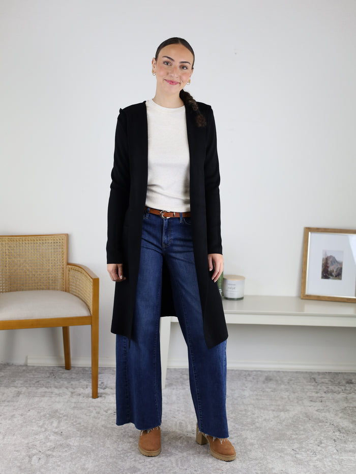 A woman stands indoors wearing the Pinch Me Long Cardigan layered over a cozy cream sweater, wide-leg blue jeans, and brown shoes. She is smiling slightly. Behind her are a wooden chair, a low white table, a candle, and a framed picture—creating the perfect setting for effortless, cozy layering.